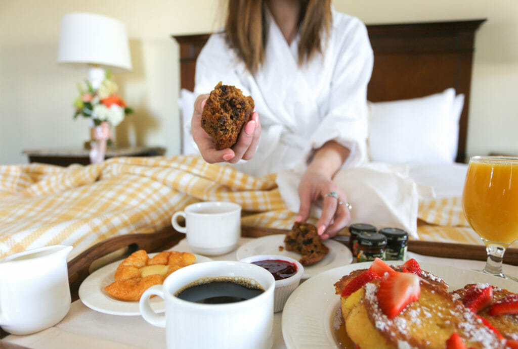 In-Room Dining tray of strawberry french toast, pastries, coffee, and orange juice on a guest room bed as woman hands her bran muffin towards the viewer.