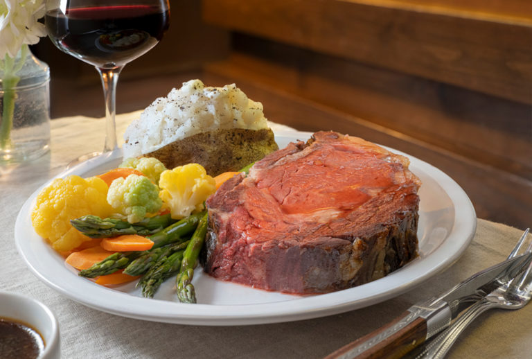 Close up of prime rib steak on a plate with mixed vegetables and a baked potato with a glass of red wine behind it.