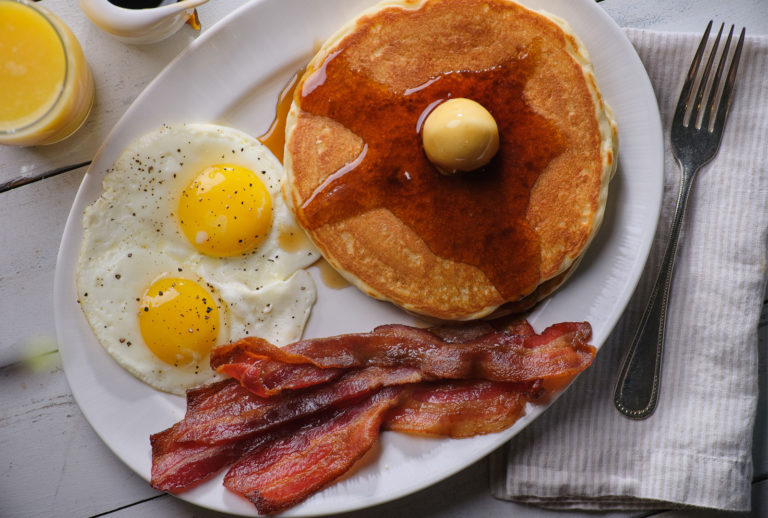 Close up of pancakes with butter and syrup, fried eggs, and bacon on a plate with a fork on the right side.